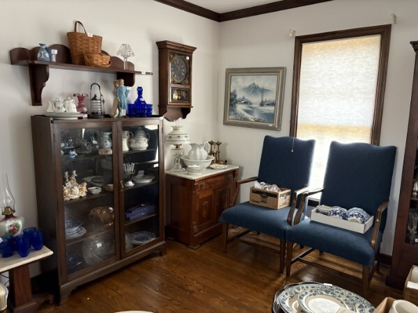Antique wooden china cabinet and marble-top washstand with blue and white china, cobalt glass, wall clock, framed art, and two blue upholstered armchairs in a wood-floor room