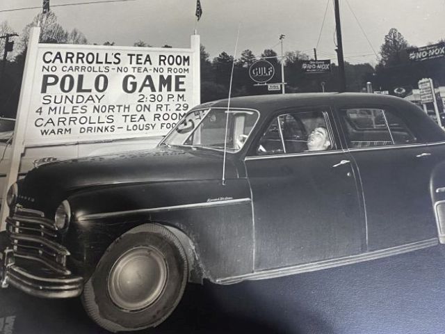 Black and white photograph from 1955 showing a dark vintage sedan parked beside a large roadside sign for Carroll’s Tea Room in Charlottesville, Virginia. The sign also advertises a Sunday polo game and includes smaller lettering about location and refreshments. In the background are utility poles, trees, and period Gulf and No-Nox signs, giving the scene a clear mid-century roadside atmosphere.