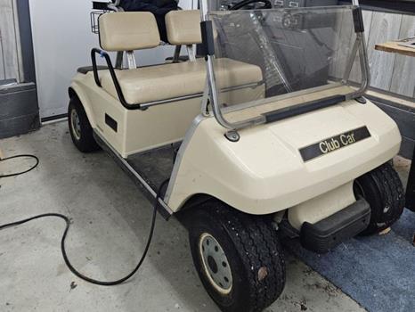 Club Car golf cart with beige body, two seats, and clear fold-down windshield parked indoors