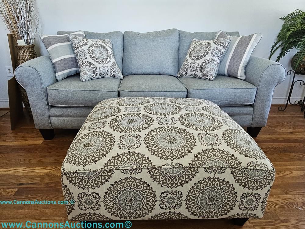 Gray three-cushion upholstered sofa with rolled arms, striped and medallion patterned throw pillows, and matching patterned upholstered ottoman.